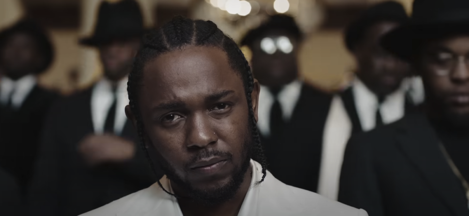 Photo of a guy with white attire and braided hair, with blurred individuals wearing suits in the background