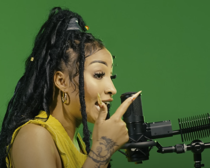 Photo of a woman with dreadlocks and long nails singing into a studio microphone, with a green background