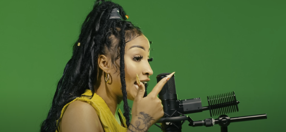 Photo of a woman with dreadlocks and long nails singing into a studio microphone, with a green background
