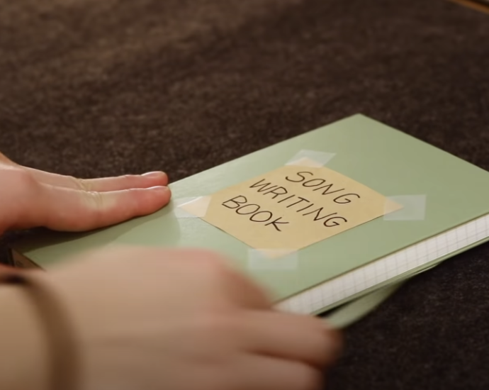 Photo of a hand holding a green notebook with a sticky note indicating 'songwriting book’