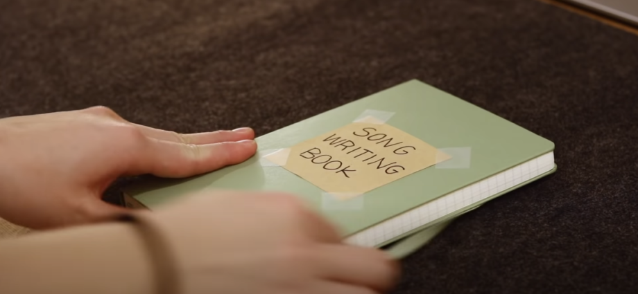 Photo of a hand holding a green notebook with a sticky note indicating 'songwriting book’