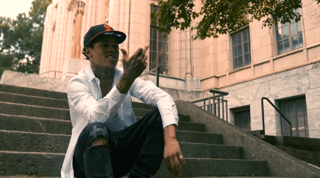 30 deep grimeyy in dark pants, hat, and white shirt sitting on the stair near building