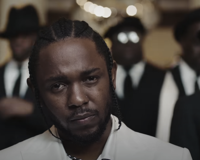 Photo of a guy with white attire and braided hair, with blurred individuals wearing suits in the background
